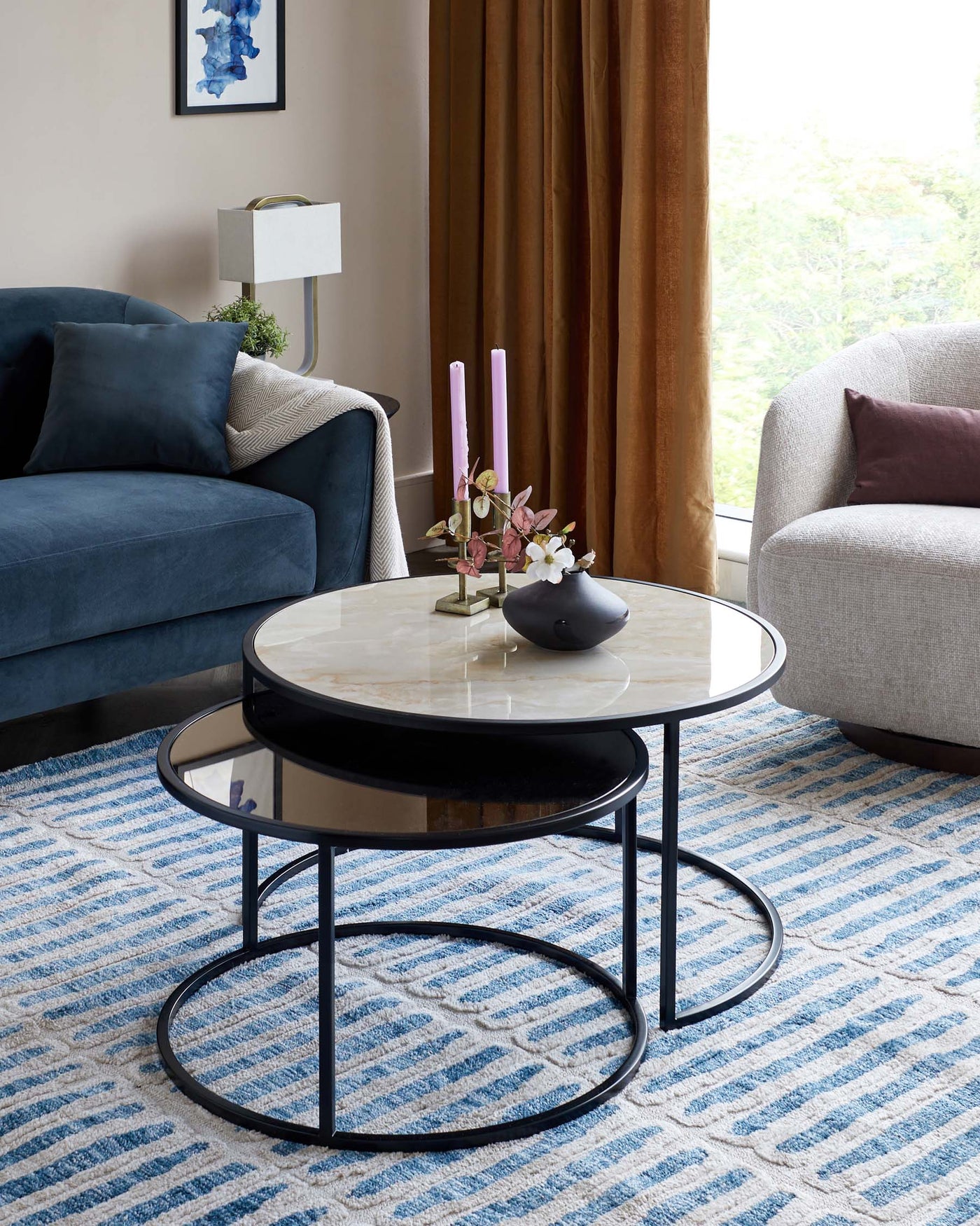A set of two modern nesting coffee tables with circular tabletops; the larger table features a white marble surface, and the smaller table has a glossy black finish, both supported by thin black metal frames. In the background, a plush navy blue upholstered sofa with a textured throw and a light grey fabric chair with a deep red cushion can be seen.