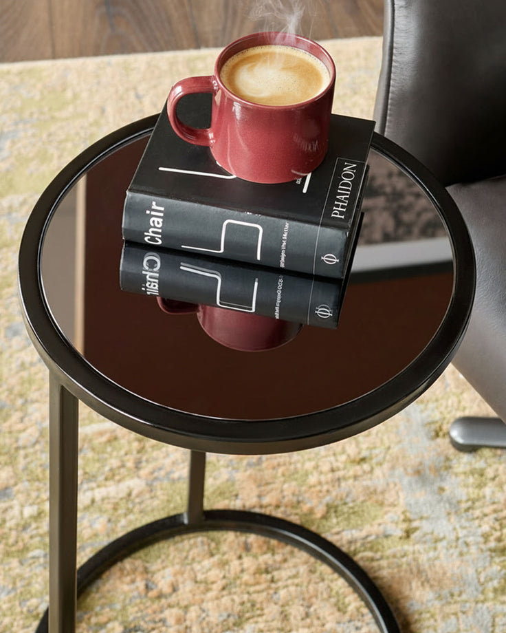 Black round side table with smoked mirror top, holding a red mug of coffee and stacked black books.