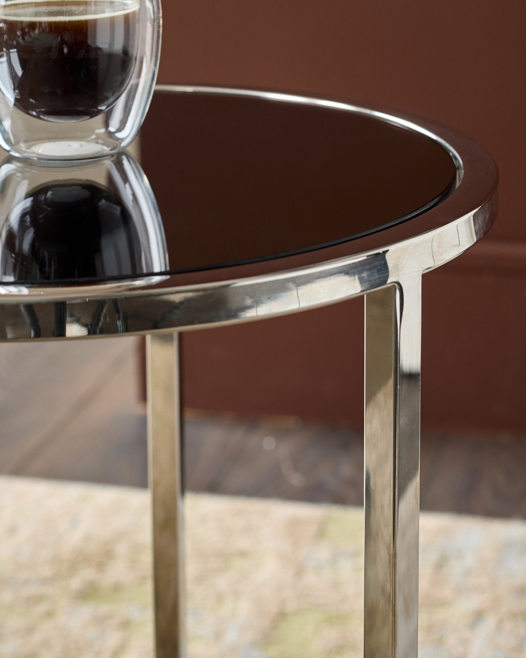 Smoked mirror tray table with brushed stainless steel frame, showing round top and angled leg detail against brown background.