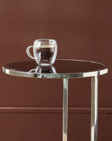Thea smoked mirror tray table with stainless steel frame holding a double-wall glass mug against brown background.
