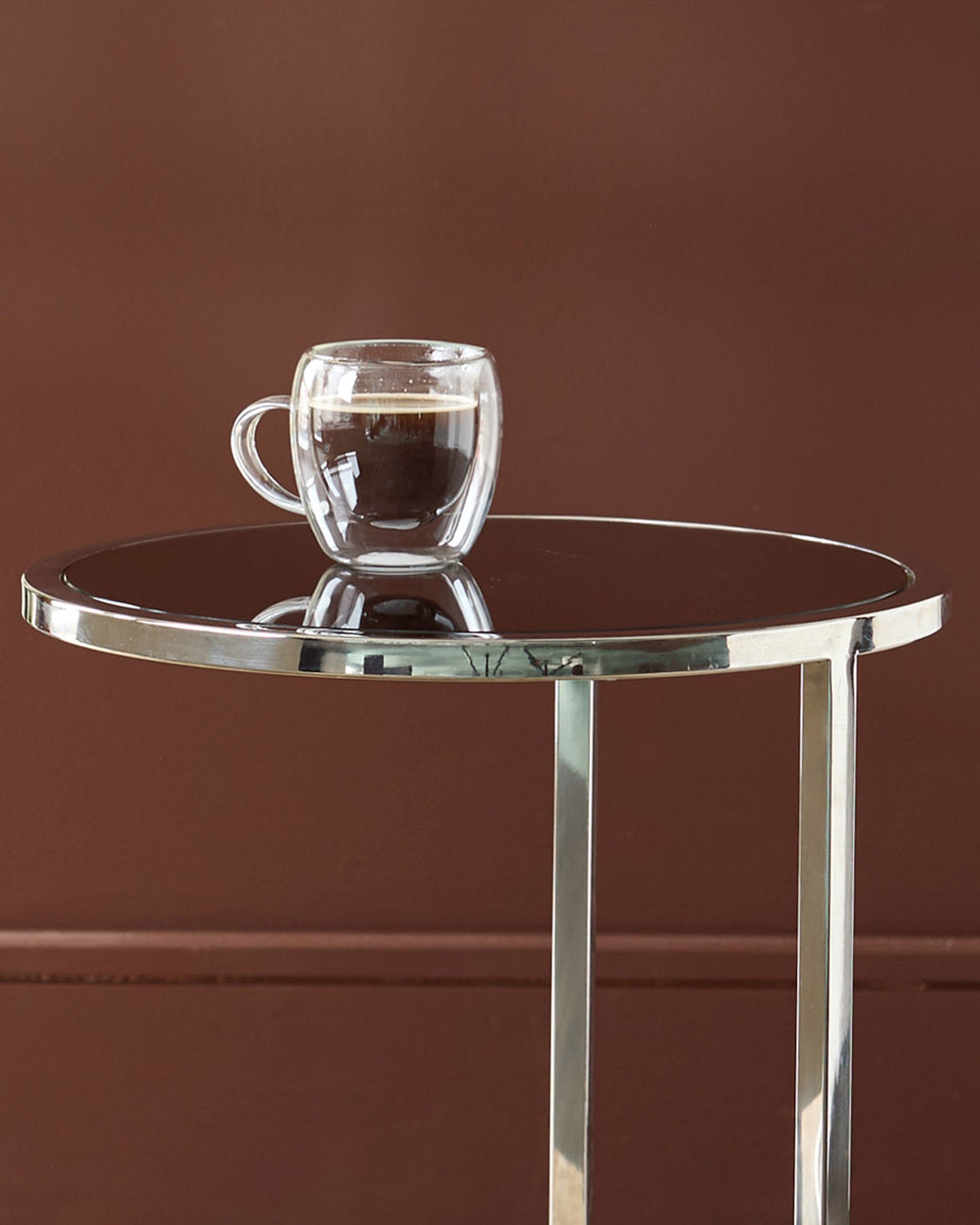 Thea smoked mirror tray table with stainless steel frame holding a double-wall glass mug against brown background.