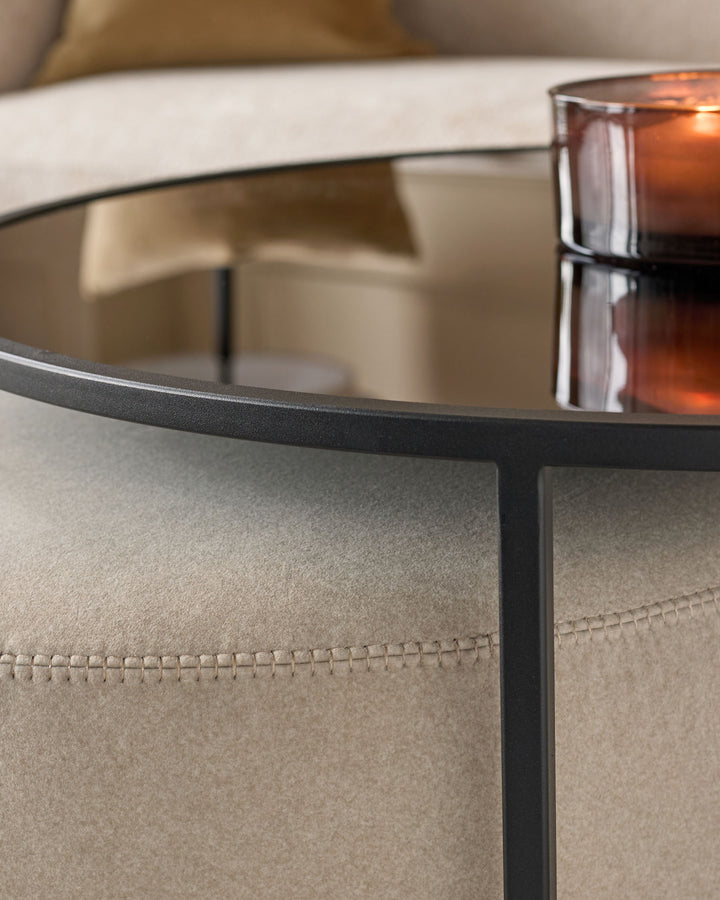 Black round coffee table with mirrored top and cream pouffe with stitched detail, styled with amber glass candle.