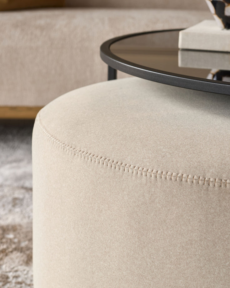 Cream upholstered cylindrical pouf with visible stitching detail, paired with a black round coffee table in a living room setting.
