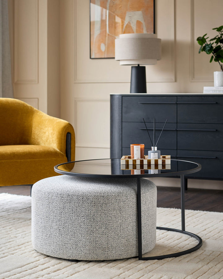 Thea black round coffee table with grey upholstered pouffe, styled in modern living room with yellow chair and dark dresser.