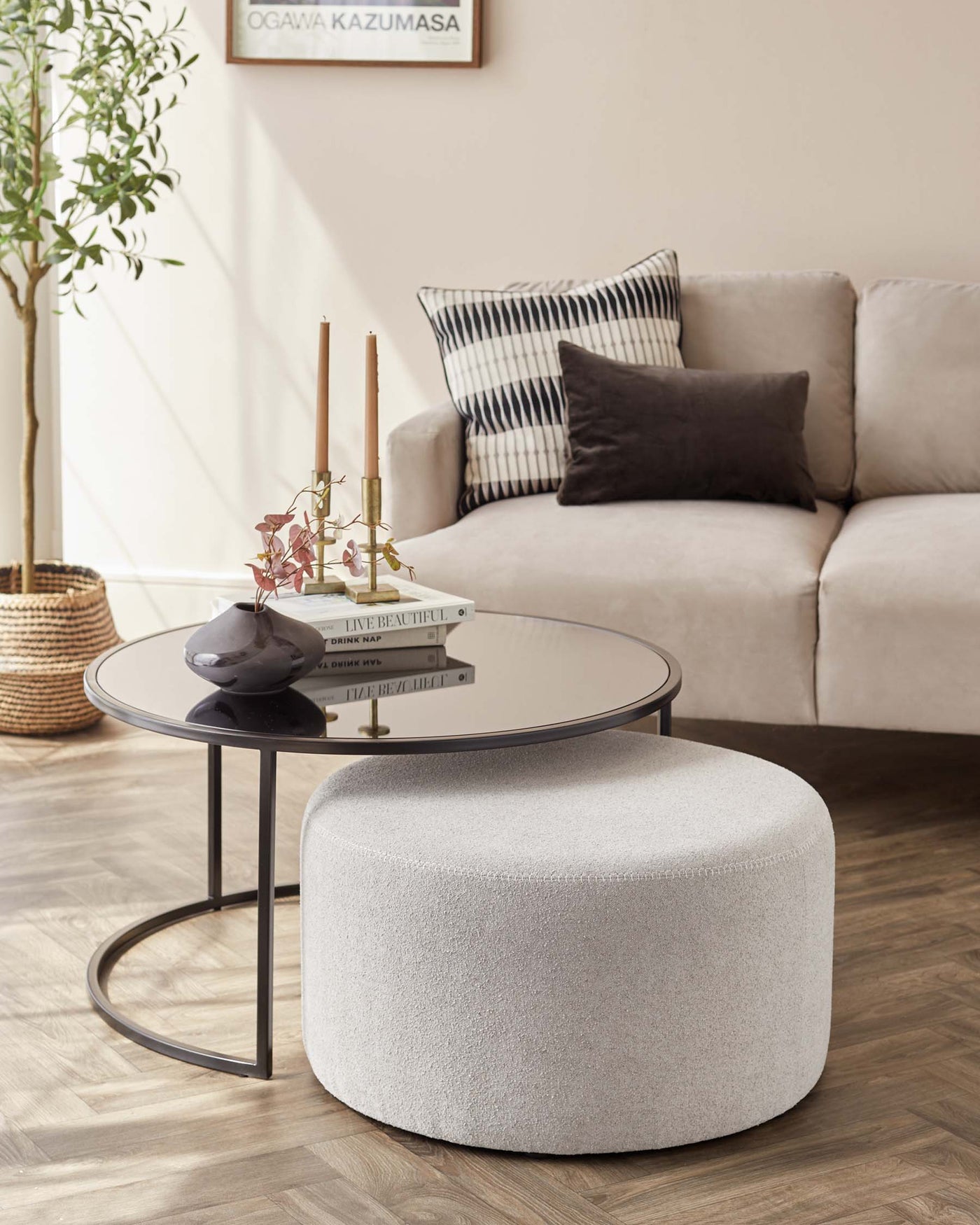 Contemporary living room featuring a light beige corner sofa with assorted throw pillows, a round glass-topped coffee table with a sleek black metal frame, and a round, fabric-upholstered ottoman in light grey. The setup is complemented by soft natural light and indoor greenery.