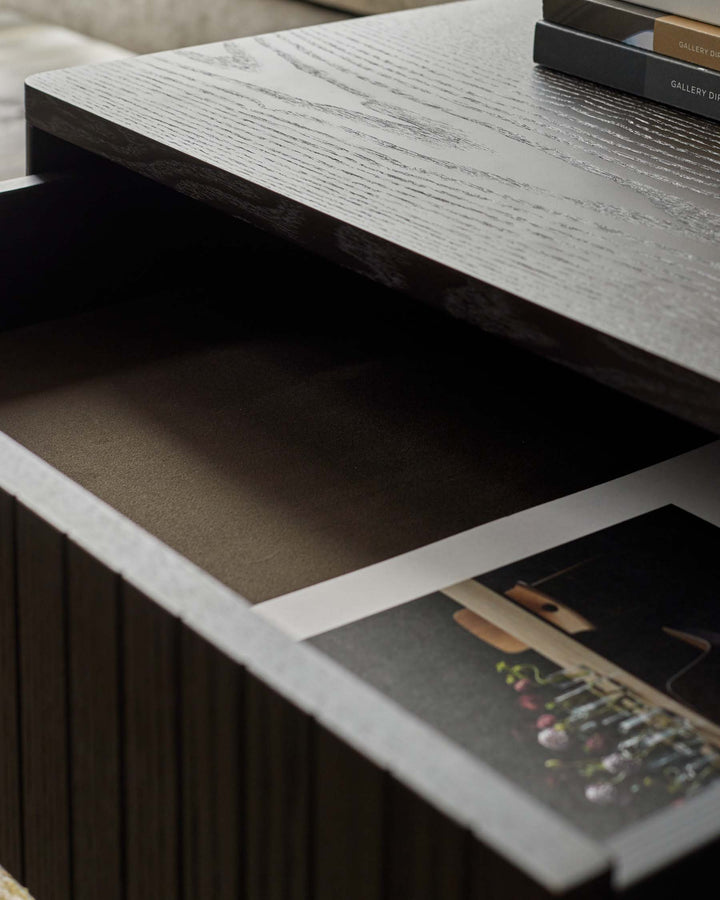 Close up of a dark wooden side table with an open drawer, lined with a soft material. A book sits inside.