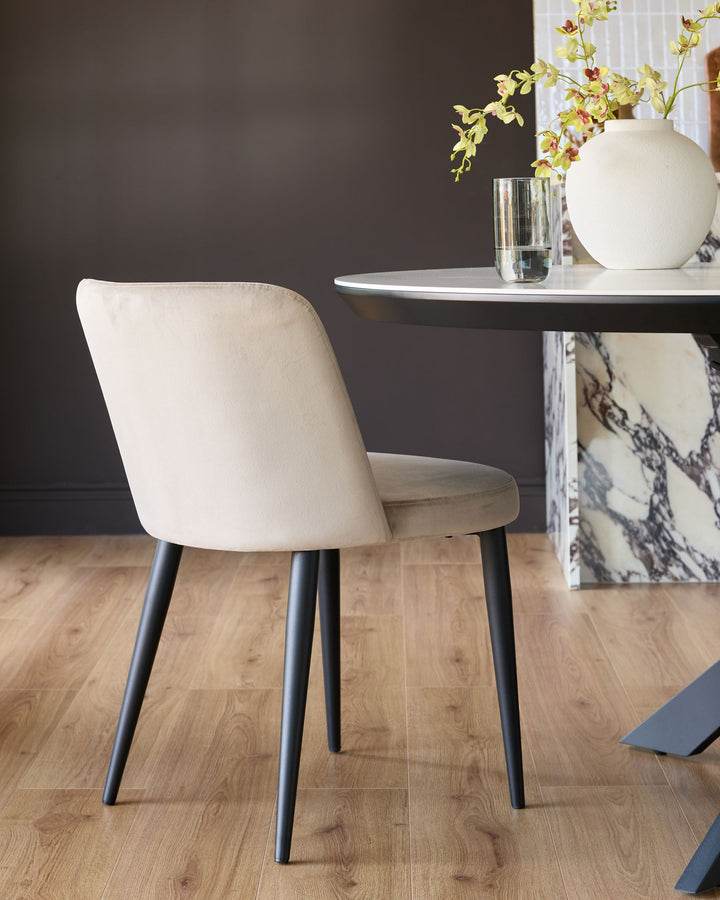 Champagne velvet dining chair with curved backrest and black tapered legs, shown from rear angle in a styled room setting.