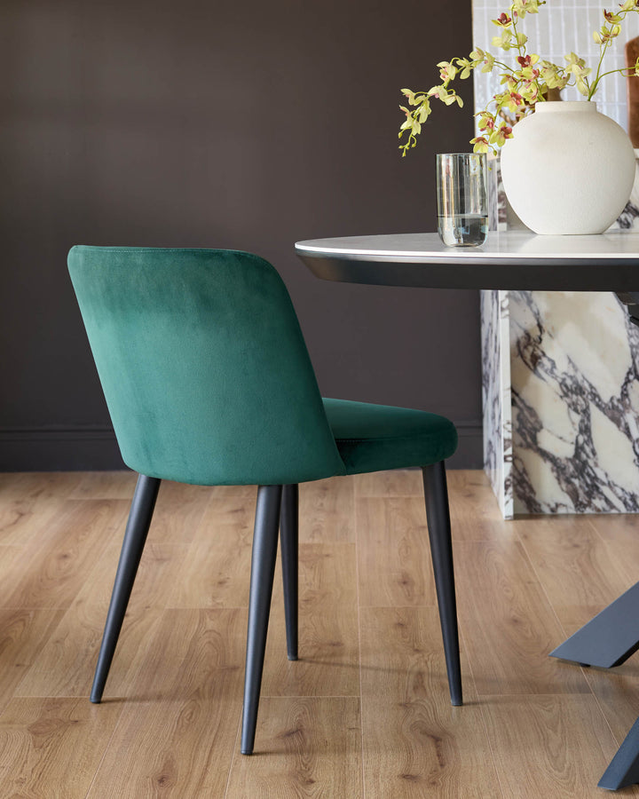Trinny velvet dining chair in dark green with black tapered legs, shown from rear angle at a white round table with marble base.