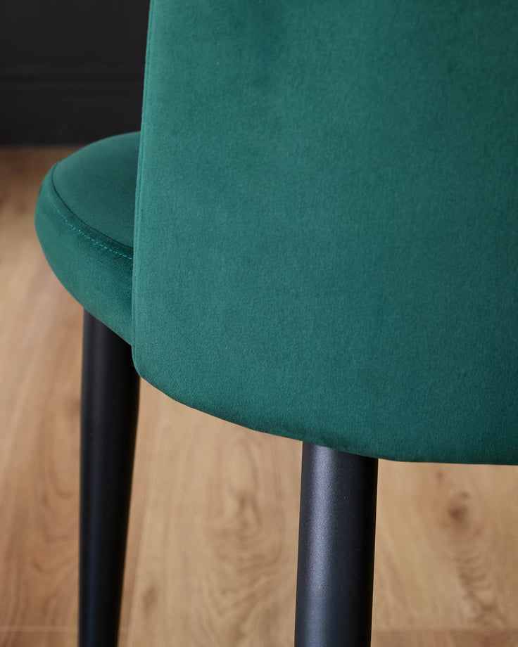Trinny velvet dining chair in dark green, angled side view showing curved backrest and black legs on wooden floor.