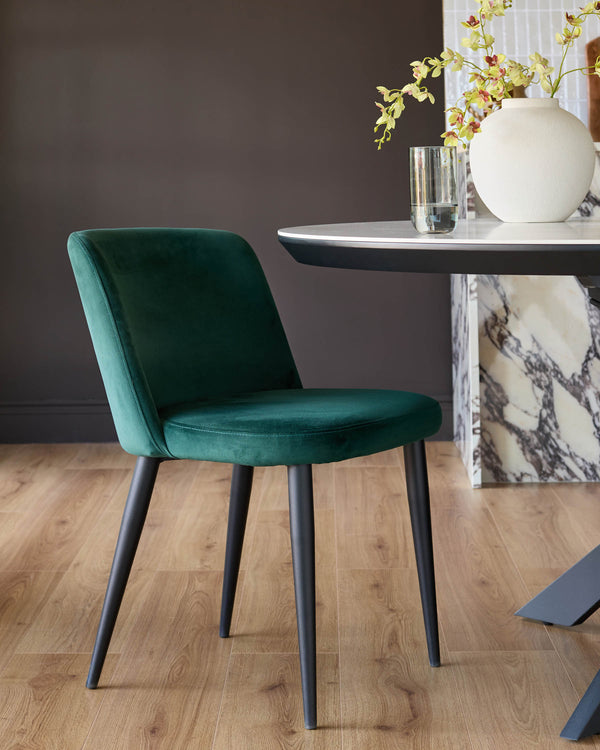 Trinny velvet dining chair in dark green with black tapered legs, shown in a styled room setting beside a marble table.