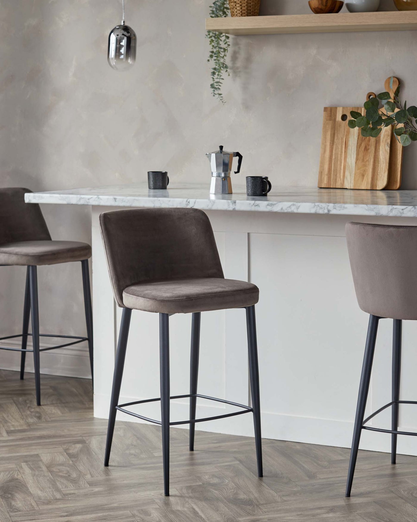 Modern kitchen interior with a pair of elegant upholstered bar stools featuring grey fabric seats and black metal legs, alongside a white marble countertop bar.