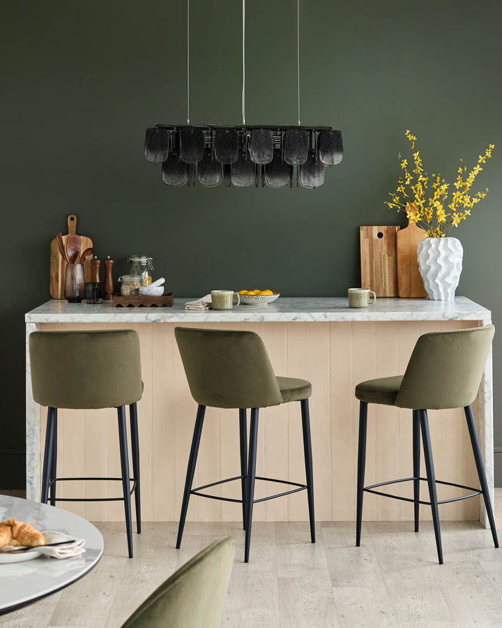 A kitchen bar with three olive green velvet stools. Marble counter with various kitchen accessories and floral arrangement.