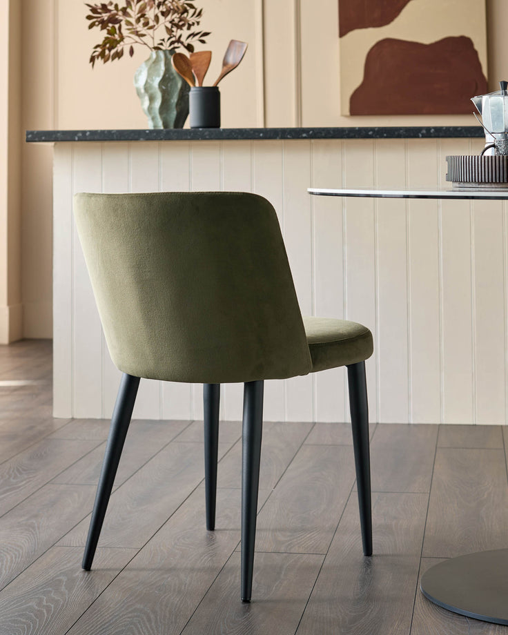 An olive green chair with black legs is positioned next to a round table in a bright room.