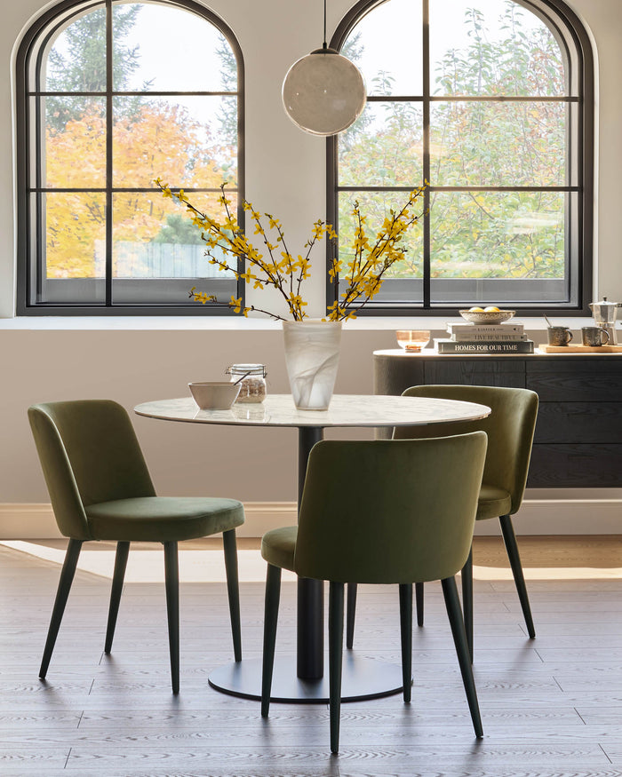 Dining area featuring a round marble table, green velvet chairs, and a black sideboard. Light and airy ambiance.