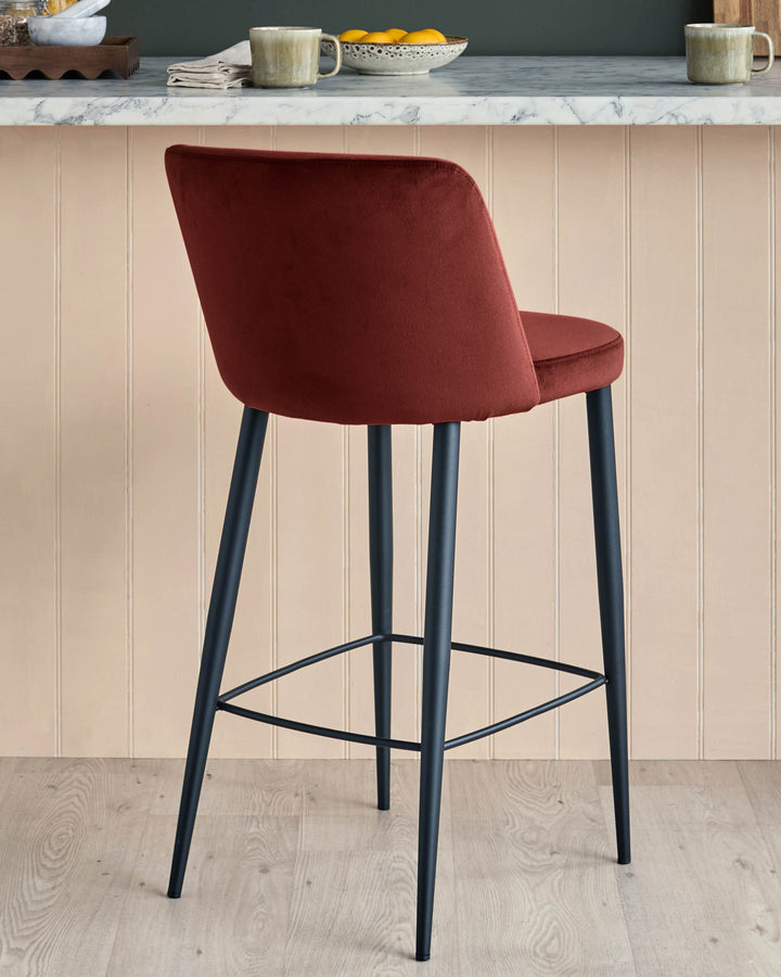 A modern bar stool in a warm red velvet, with sleek black legs and footrest, sits in a kitchen setting.