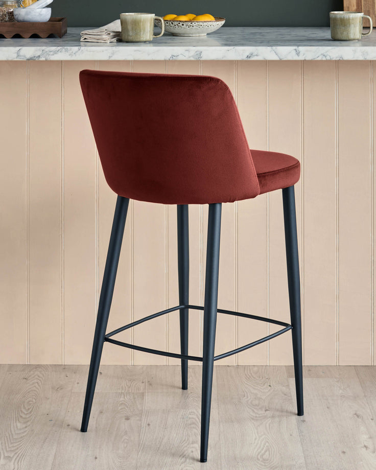 A modern bar stool in a warm red velvet, with sleek black legs and footrest, sits in a kitchen setting.