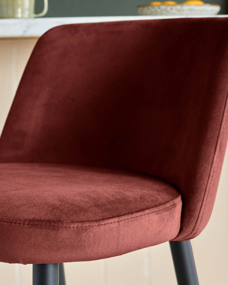 Close-up of a stylish, modern velvet chair in a rich, earthy red colour with sleek black legs. A luxurious seating option.