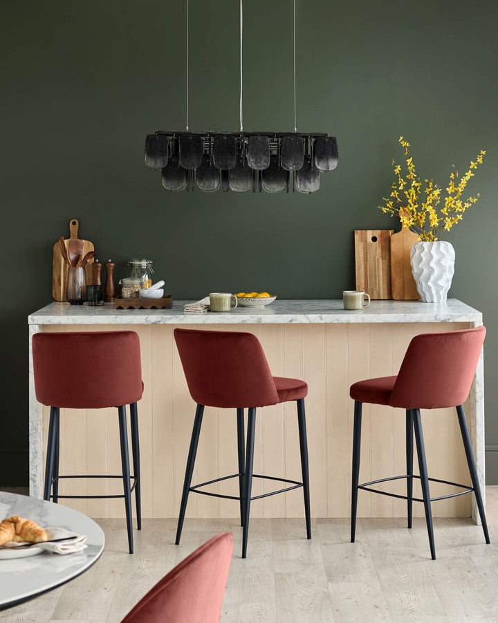 A kitchen counter with three red velvet bar stools, black legs and marble top, and overhead hanging light fixture.