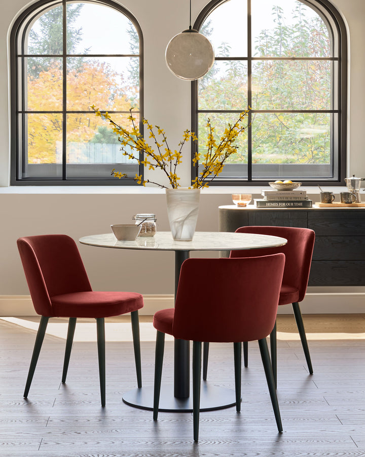 A round marble table is surrounded by three stylish red velvet chairs with dark legs. Modern dining area.