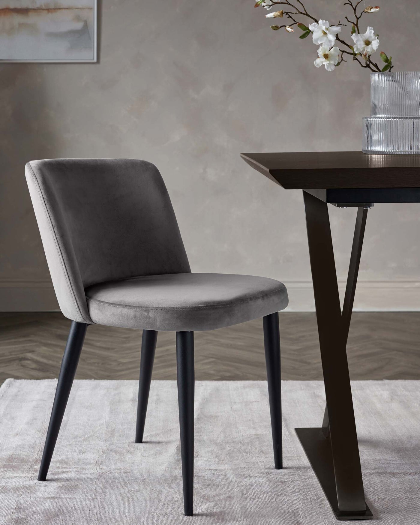 Elegant modern furniture featuring a dining chair with a curved back design and plush grey velvet upholstery, supported by four sleek, tapered black metal legs. Adjacent is a contemporary dark wooden table with an angular base showcasing a minimalist aesthetic. A light area rug under the chair complements the set.