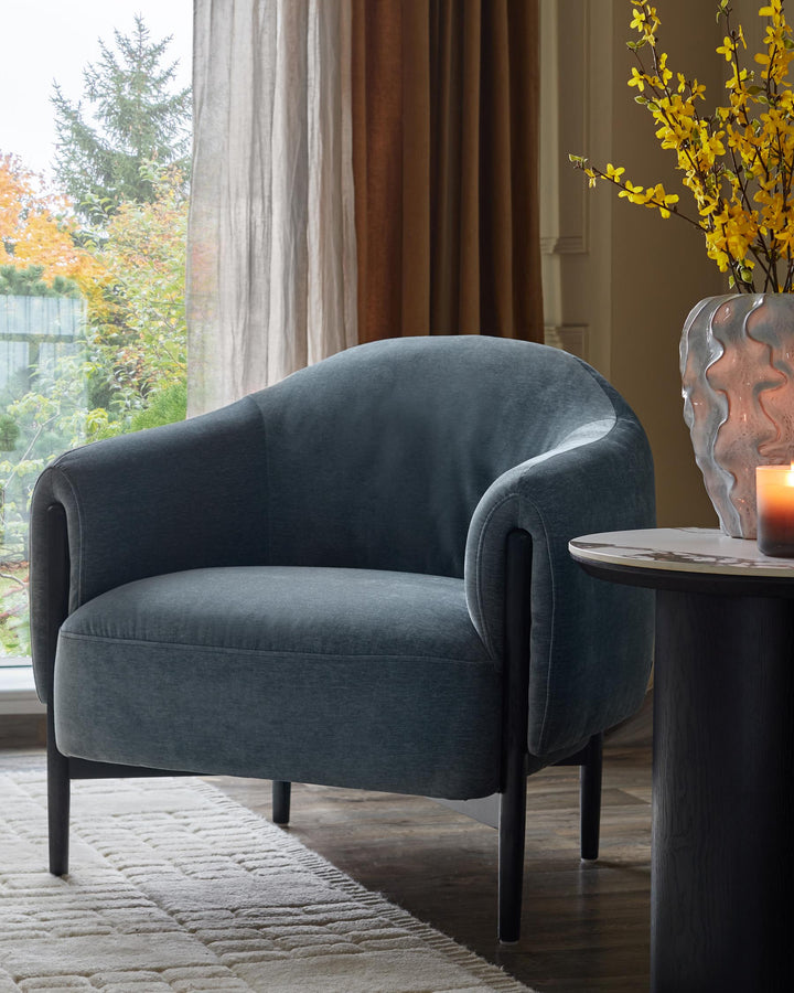 A blue armchair sits on a cream rug next to a black side table with a marble top, beside a window.
