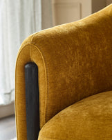 Close-up of a stylish mustard velvet chair with a curved back and black wood arm support. Luxurious and comfortable.