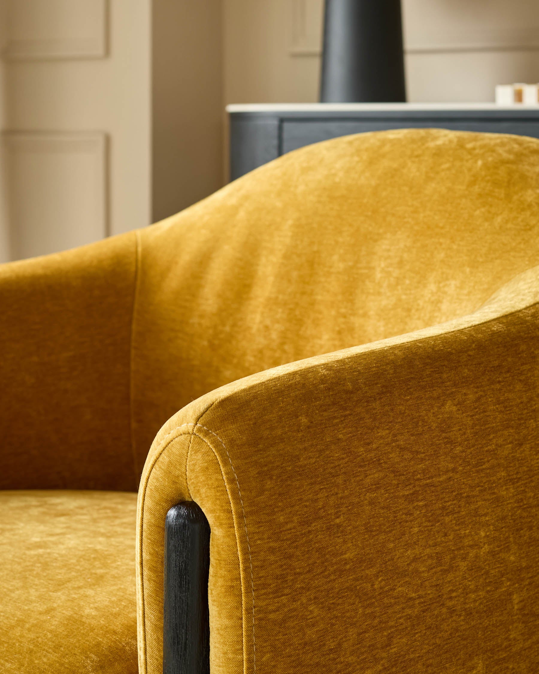 Close up of a mustard yellow velvet armchair, with a dark wooden arm detail and light-coloured walls in the background.