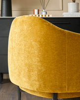 Close-up of a mustard yellow velvet armchair with black legs, in front of a black cabinet with accessories.