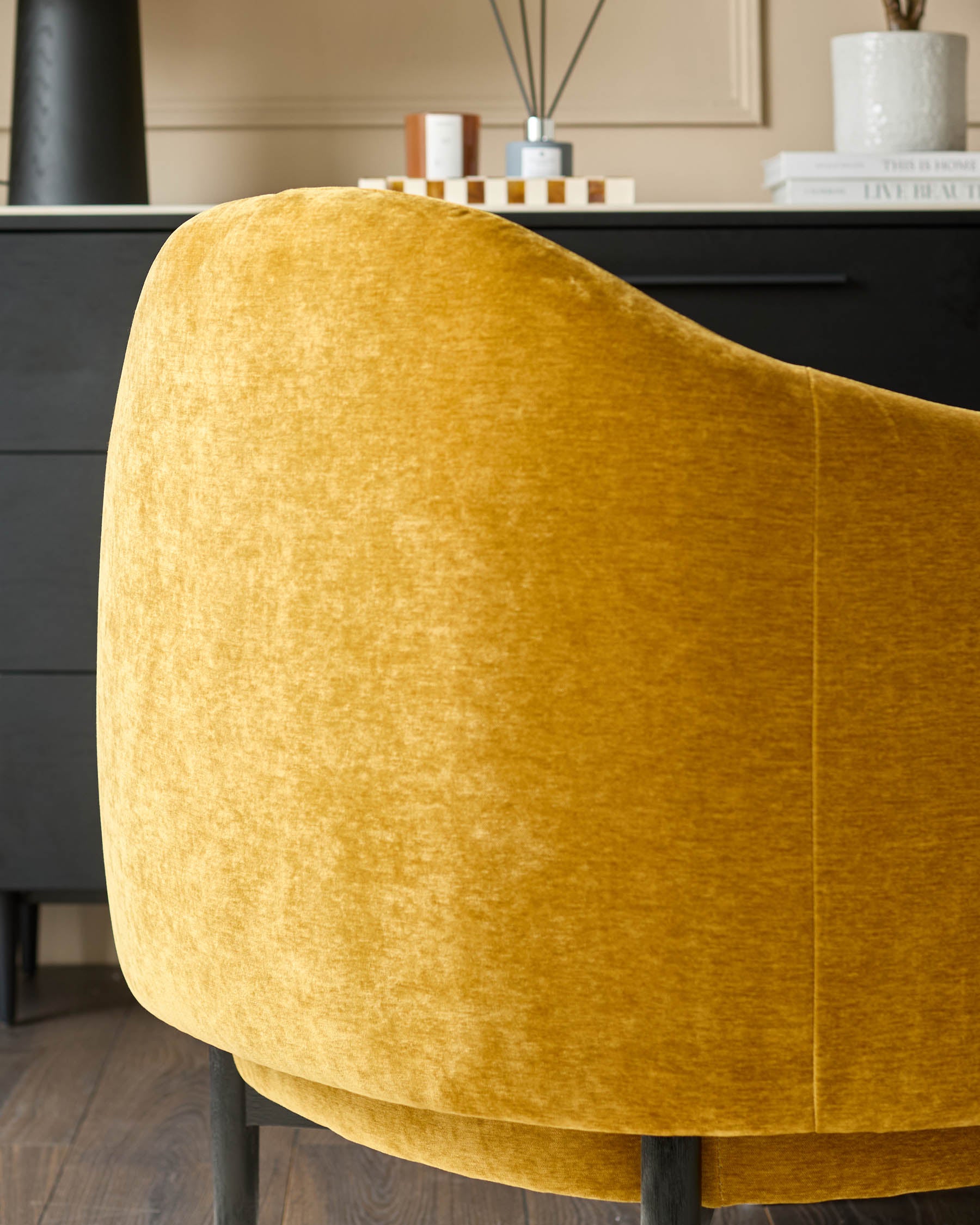 Close-up of a mustard yellow velvet armchair with black legs, in front of a black cabinet with accessories.