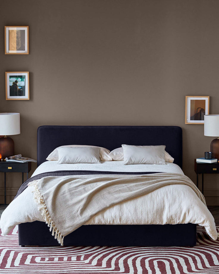 Bedroom with a navy upholstered bed, bedside tables with lamps, artwork on the walls, and a patterned rug.