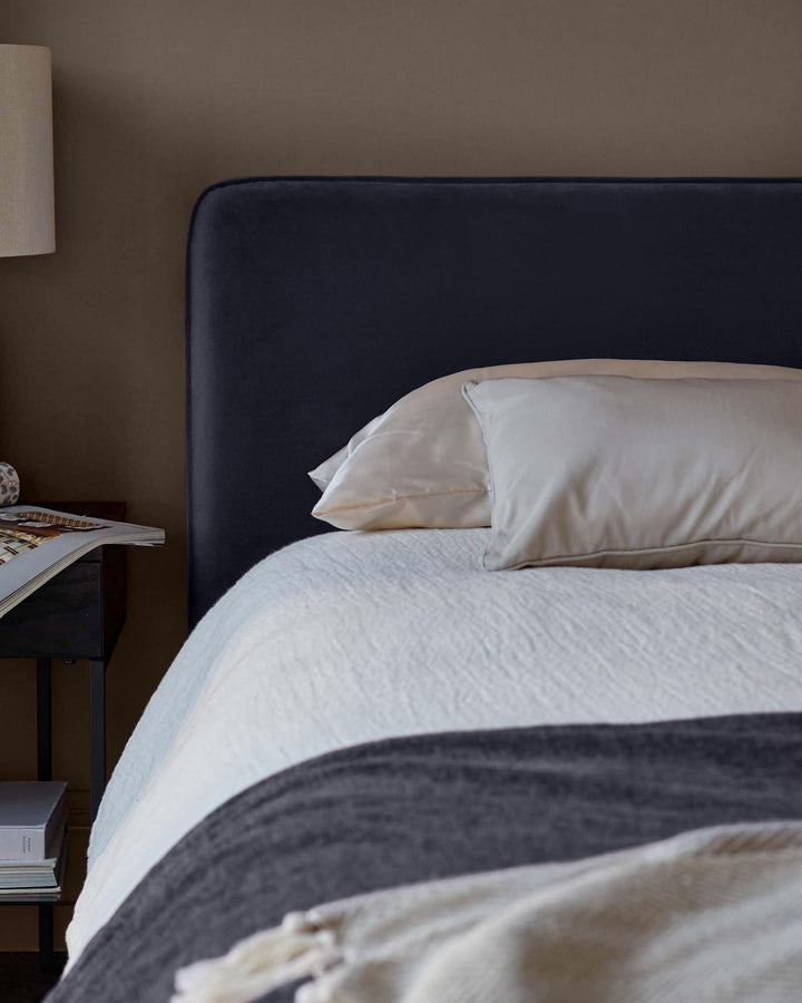 Upholstered bed with headboard, pillows, bedding and throw; bedside table with lamp and books.