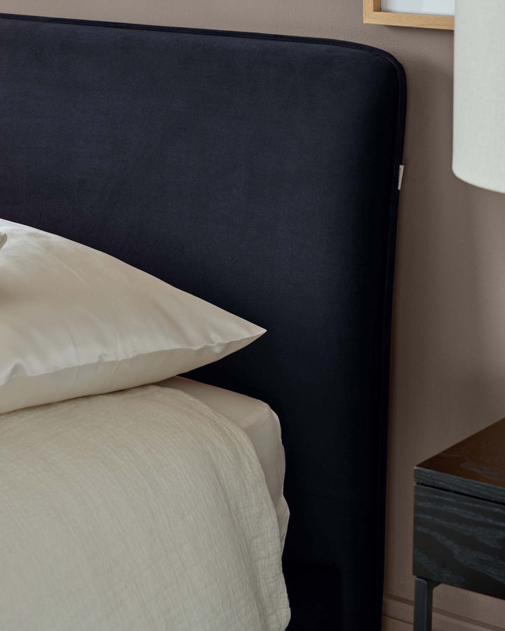 A stylish bed with a dark headboard, dressed with a plush white pillow and light textured throw, next to a dark wooden side table.