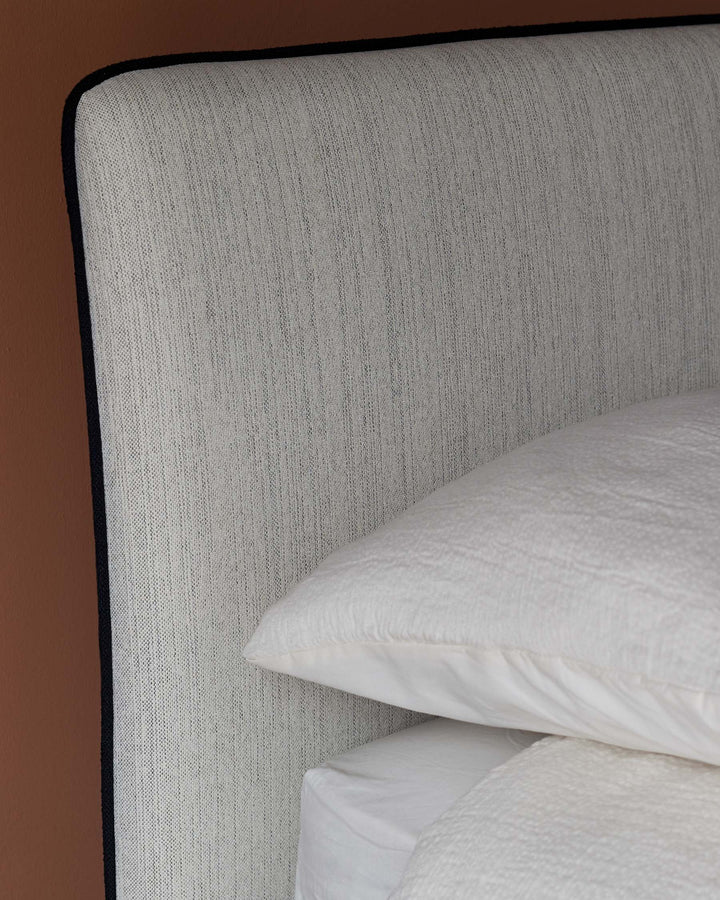 Modern bed headboard with a textured light grey fabric and contrasting black trim, paired with white pillows and bedding.