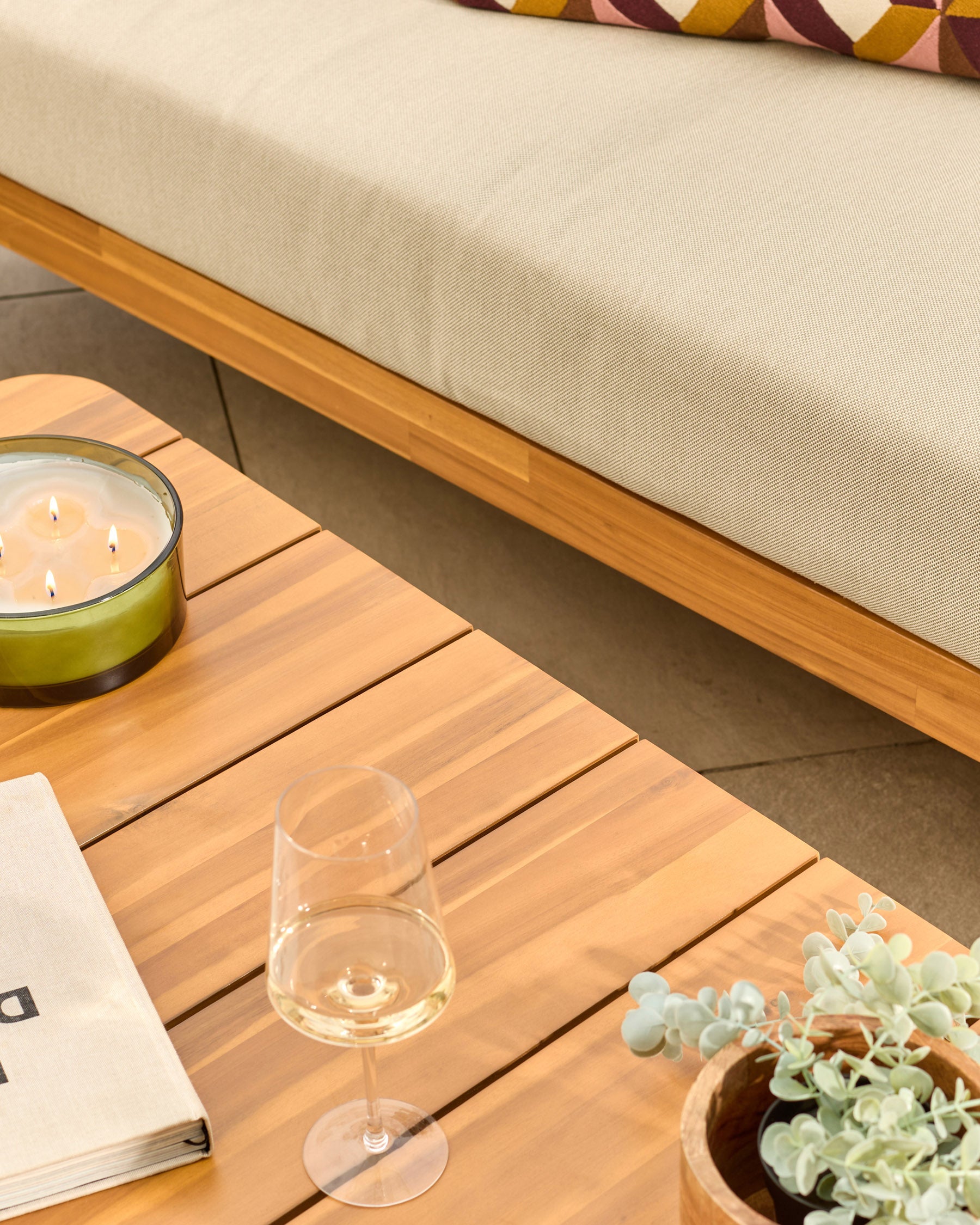 Natural wood garden sofa set with cream cushions, wooden coffee table, wine glass, candle, and potted plant decoration.