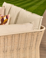 Natural woven rattan basket with cream cushions and decorative pillow, outdoor setting with green lawn background.