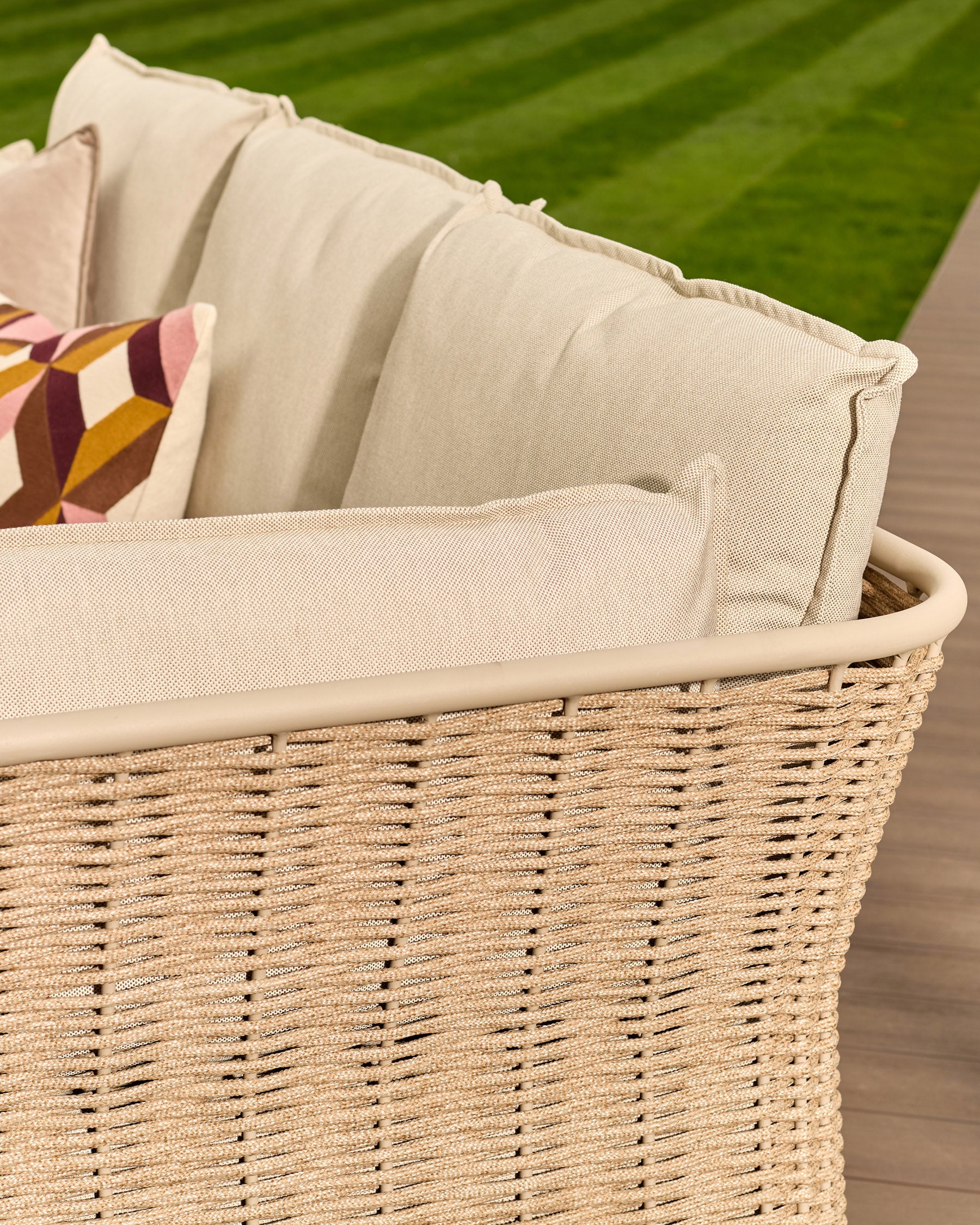 Natural woven rattan basket with cream cushions and decorative pillow, outdoor setting with green lawn background.