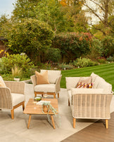 Tuscany garden set with natural wicker armchairs, 3-seater sofa, and wooden coffee table on patio, styled with cream cushions.