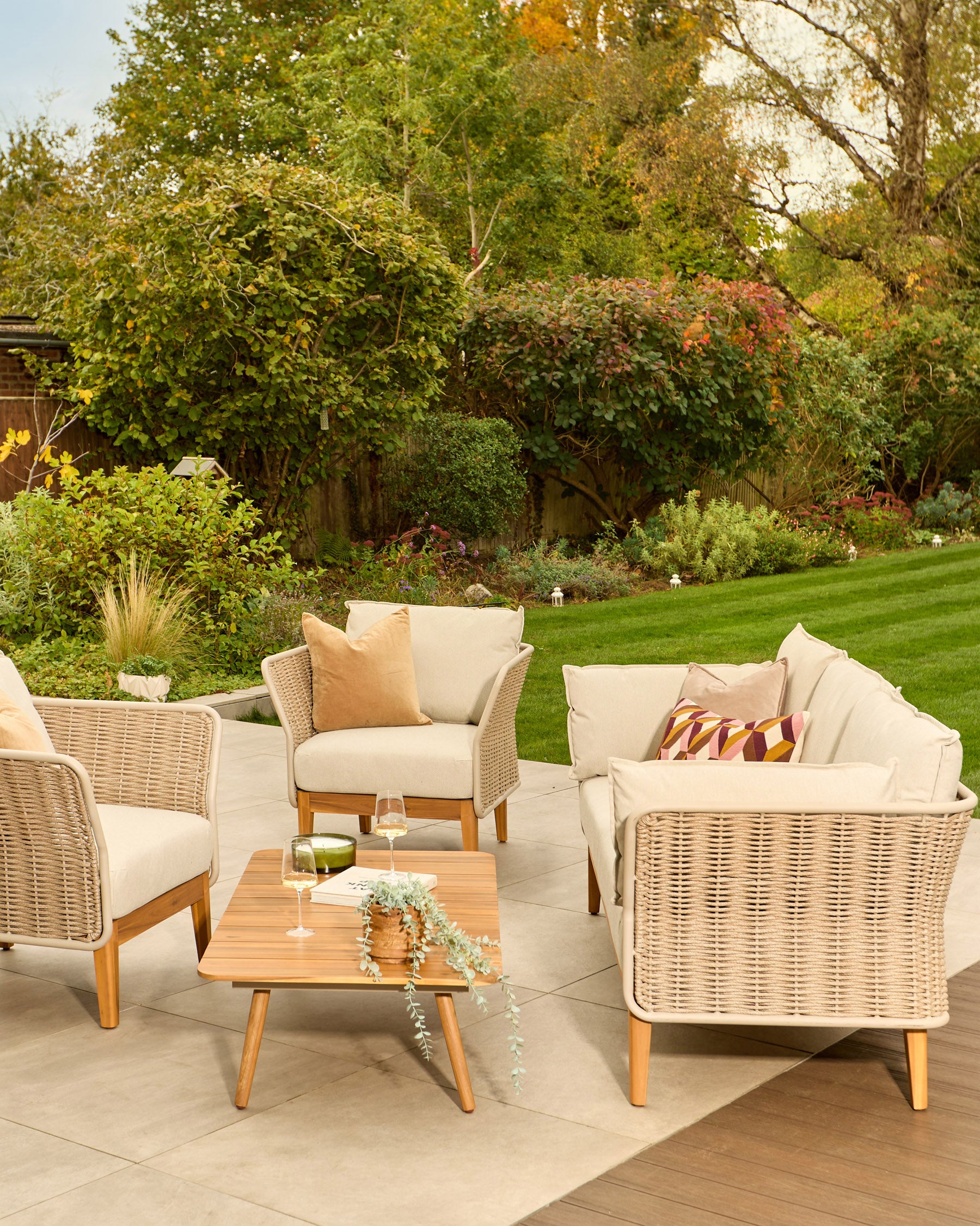 Tuscany garden set with natural wicker armchairs, 3-seater sofa, and wooden coffee table on patio, styled with cream cushions.
