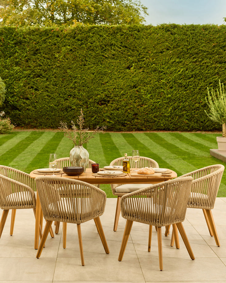 Tuscany teak wood 6-seater dining table set with woven rope chairs on a stone patio, overlooking a manicured lawn.