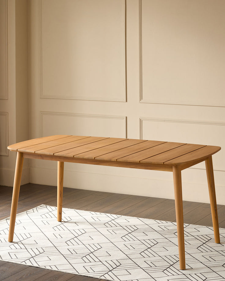Tuscany teak wood dining table with slatted top, angled legs, shown in light room with geometric patterned rug.