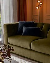 Olive green velvet 3-seater sofa with curved arms and black pillows, styled in a modern living room setting.