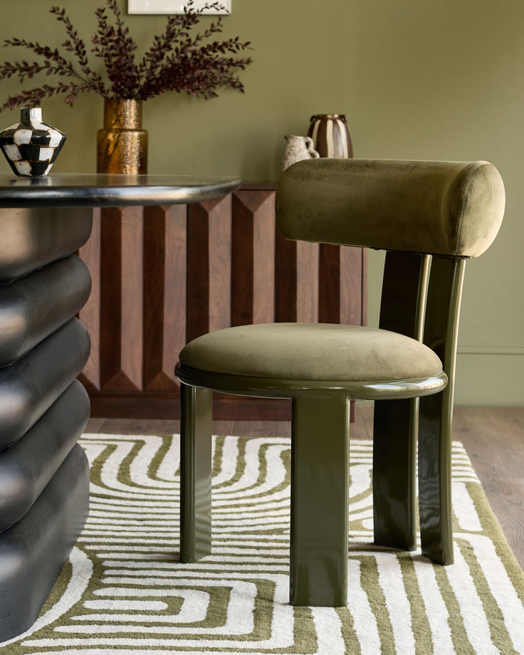 Valerie dining chair in olive green velvet with cylindrical backrest and metal legs, styled in room setting.