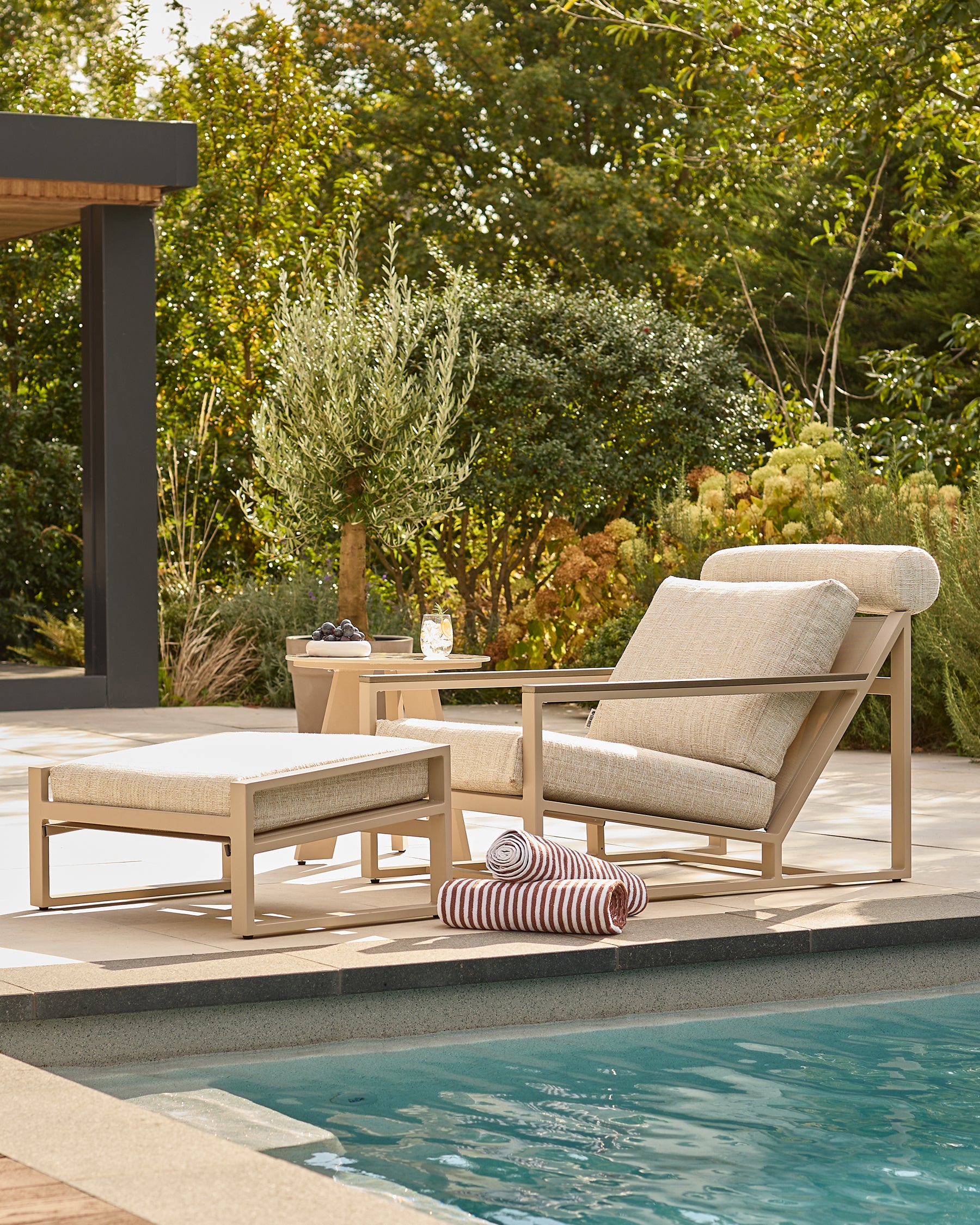 Verano lounge chair with footstool in natural wood and beige cushions, poolside setting with garden backdrop.