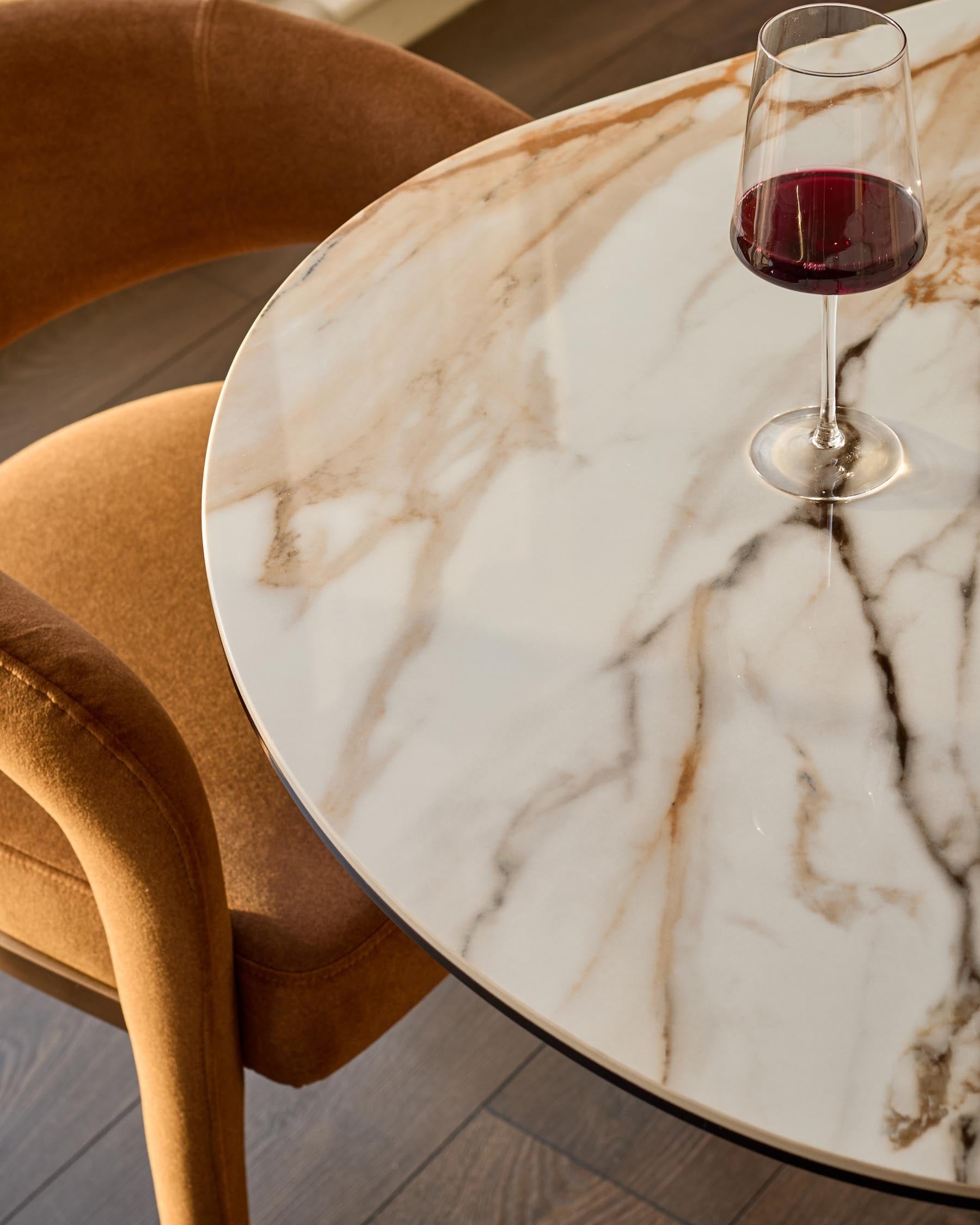 Vidal round dining table in natural ceramic with marble veining, paired with tan upholstered chair and red wine glass.