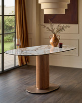 Natural ceramic oval dining table with fluted wood pedestal base and metal footrest, styled in modern dining room setting.