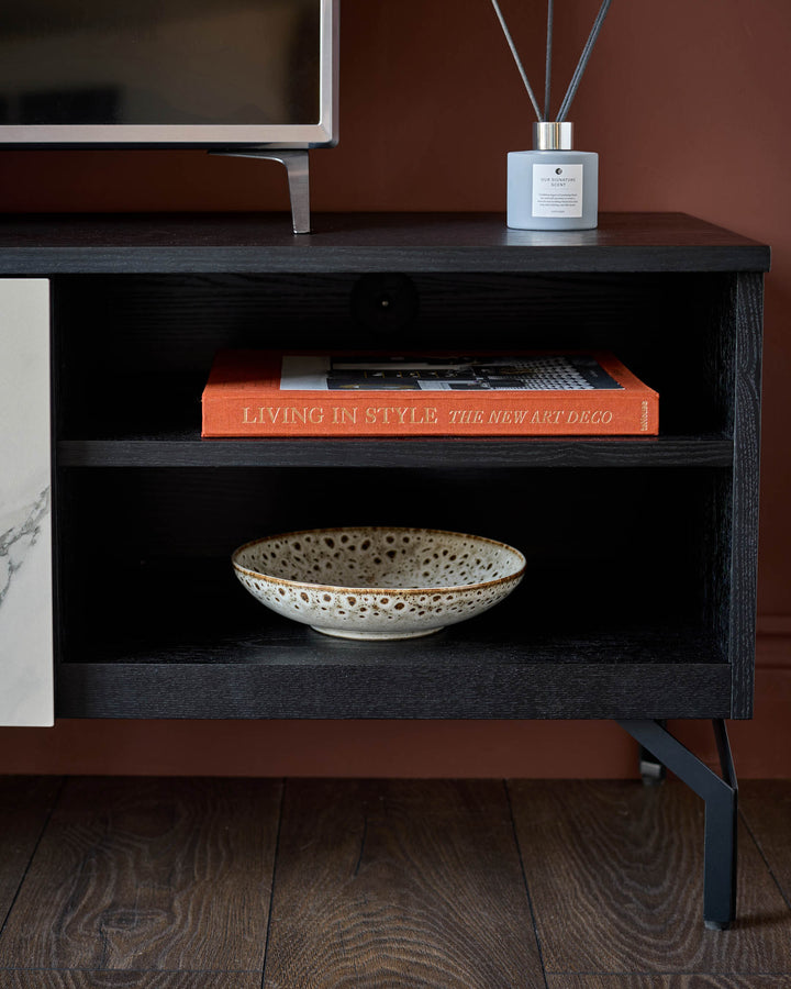 Black wood TV unit with open shelving displaying an orange book and ceramic bowl, styled with a white diffuser on top.