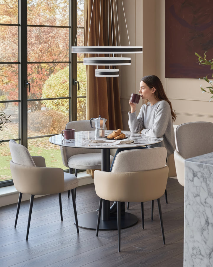 Wren dining chair in natural fabric, shown in modern dining setting with round table and autumn view through windows.