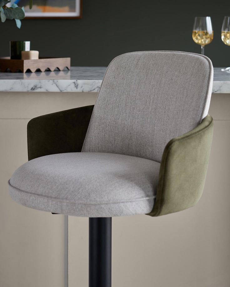 Upholstered bar stool with olive green arms and grey fabric seat. It sits on a black pole with marble worktop behind.