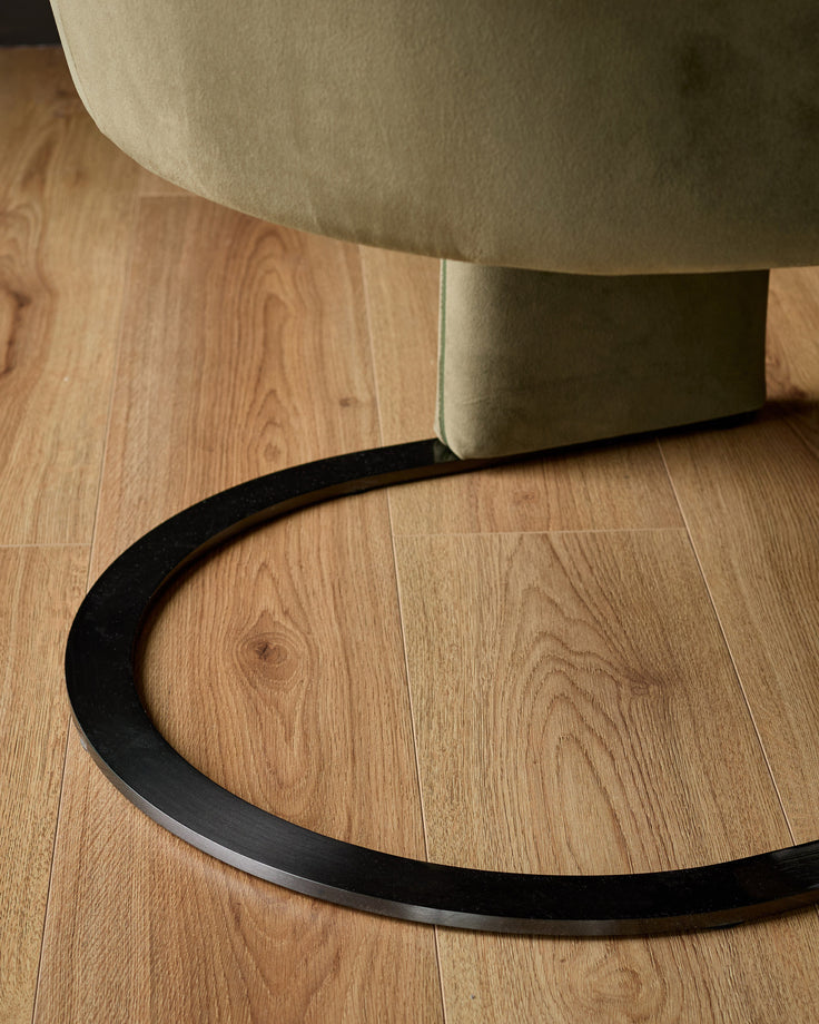 Wren cantilever dining chair in natural velvet with olive green fabric, shown from below highlighting curved black metal base on wooden floor.