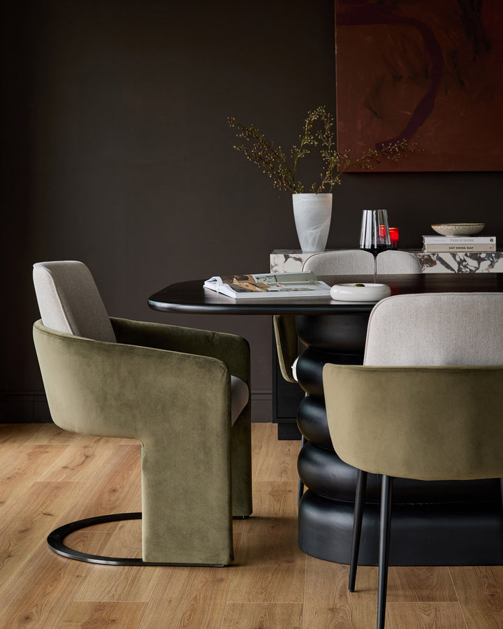 Wren cantilever dining chair in natural and olive green velvet, shown at black round table in modern dining setting.